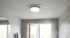 A round white bathroom ceiling fan with an integrated LED light fixture illuminated in a tiled shower area.