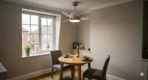 A photorealistic 4K image capturing a cozy, small-space dining area within a typical modern UK flat. A sleek, low-profile white and polished metal compact ceiling fan is centred over a round, light-wood bistro table set for two. High detail and soft, natural daylight reinforce the suitability of this smaller-scale fan.