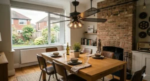A photorealistic 4K photograph of a contemporary dining room in a British home with a strong industrial-chic aesthetic. Centred above a substantial distressed light oak table is a unique industrial-style ceiling fan featuring an aged iron frame, exposed vintage filament bulbs, and wide perforated metal mesh blades. The room includes a textured brick accent wall and the large garden window consistent with the surrounding images.