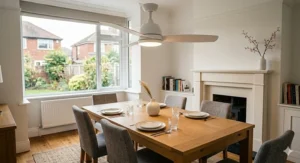 A focused, 4K photorealistic photograph showcasing a minimalist Scandinavian aesthetic. Centred under a sleek, ultra-modern minimalist ceiling fan in matte white with three sculptural white-painted wood blades is a large oak table set with clean white ceramic plates, polished silver, and crystal glasses. The background fireplace and bookshelf are simplified and decluttered.