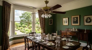 Highly detailed 4K photograph set within a grand Victorian British dining room with rich forest green walls. A substantial, ornate brass ceiling fan with polished dark mahogany blades provides a traditional touch above a dark mahogany table set with fine china and polished silver. Natural daylight illuminates the intricate textures of the coving and formal garden view.