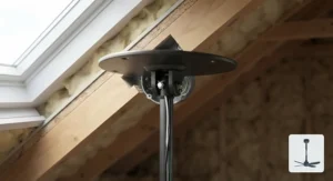A view inside the roof space showing the matt black ceiling fan downrod securely bolted to the pivoting sloped ceiling adapter. A cotter pin and safety bolt are visible, ensuring a safe connection. The surrounding yellow insulation and rafter structure are in focus.
