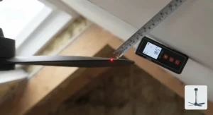 A macro photograph illustrating the measurement of the blade tip clearance for a ceiling fan on a sloped ceiling. A tape measure is extended from the tip of a matt black fan blade to the angled white plasterboard ceiling, ensuring sufficient distance is maintained.