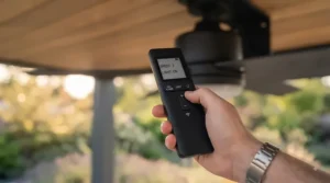 A hand holding a smart remote control adjusting the speed settings of an outdoor ceiling fan on a covered terrace.